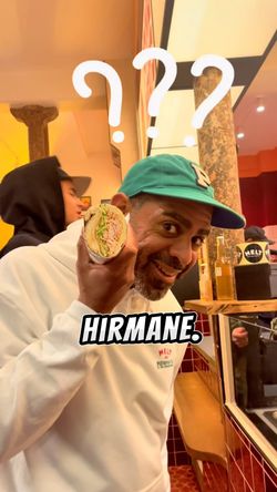 Un client souriant tient un sandwich garni de viandes variées dans un cadre de restaurant animé.