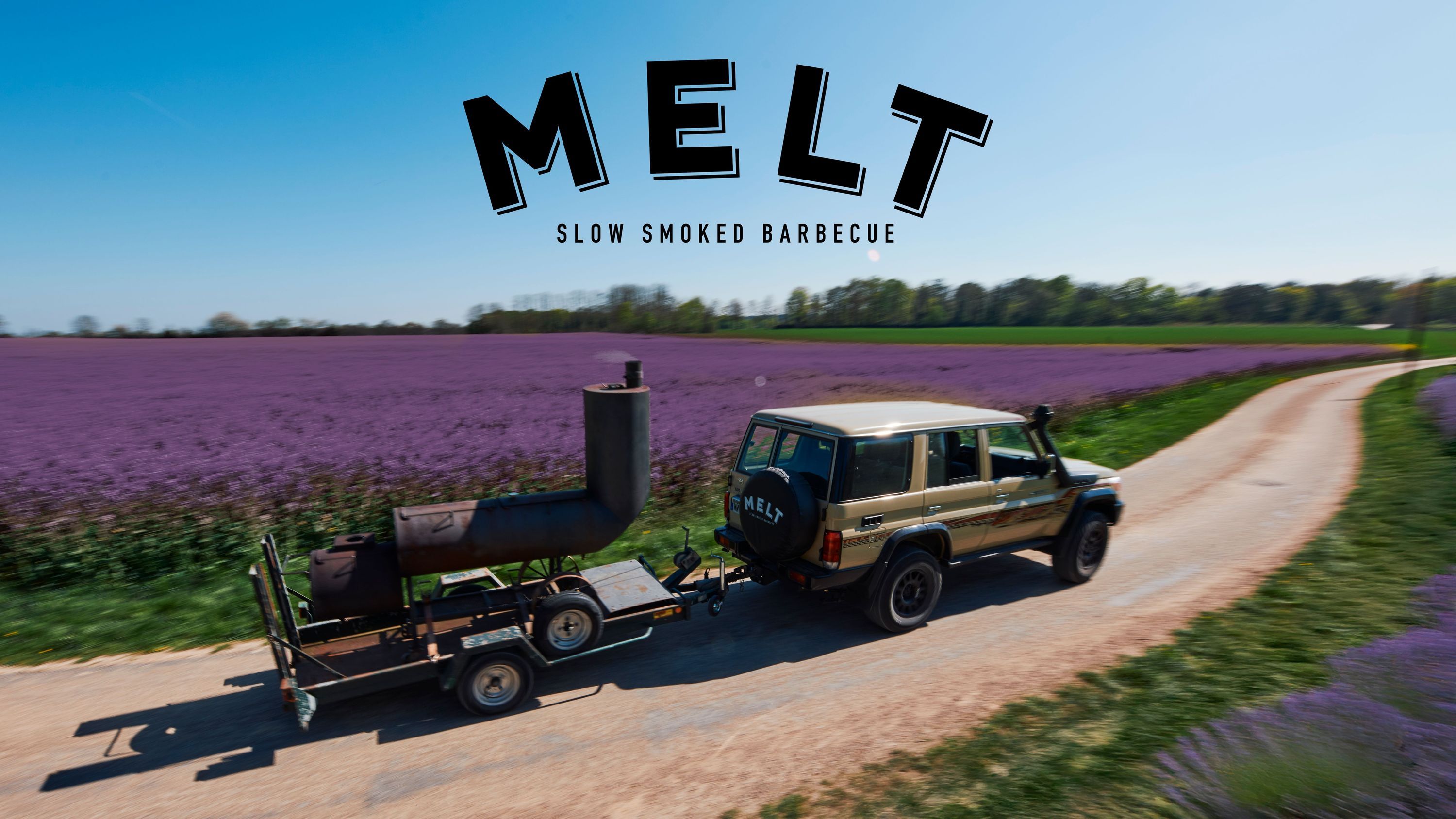 A trailer equipped with a smoker is being towed by a vehicle along a scenic road surrounded by blooming fields.