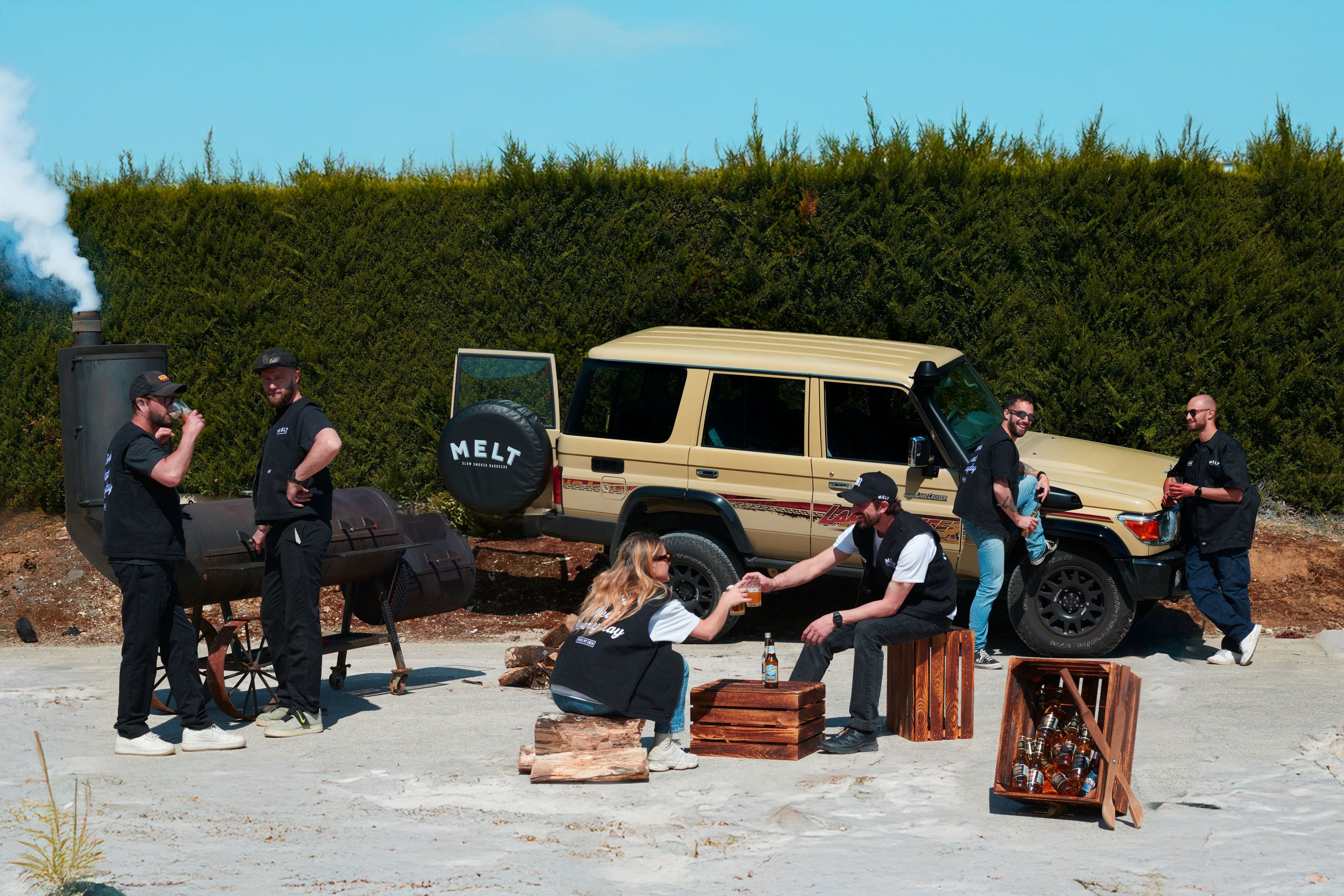 Un groupe de personnes à l'extérieur, avec un véhicule tout-terrain vintage et un fumoir à barbecue.