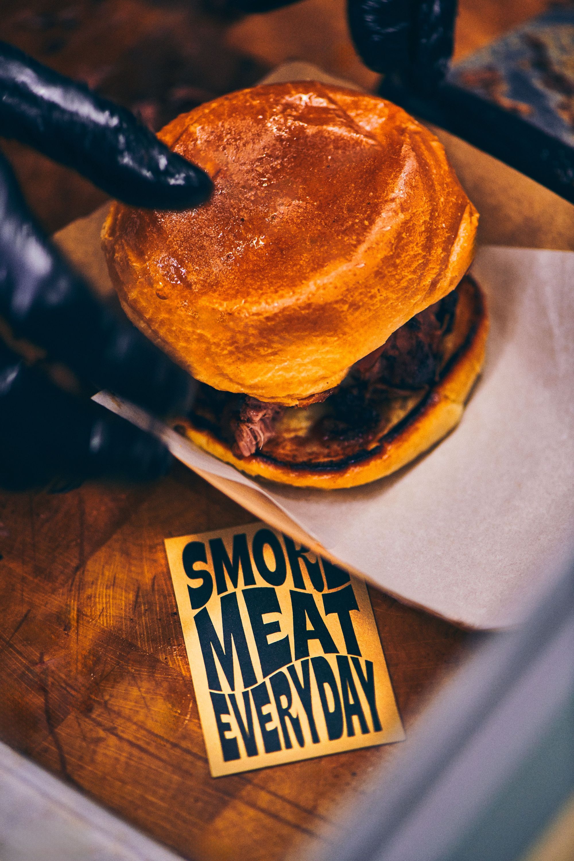 A close-up of a brisket sandwich featuring a golden bun, accompanied by a card that reads "Smoked Meat Everyday."