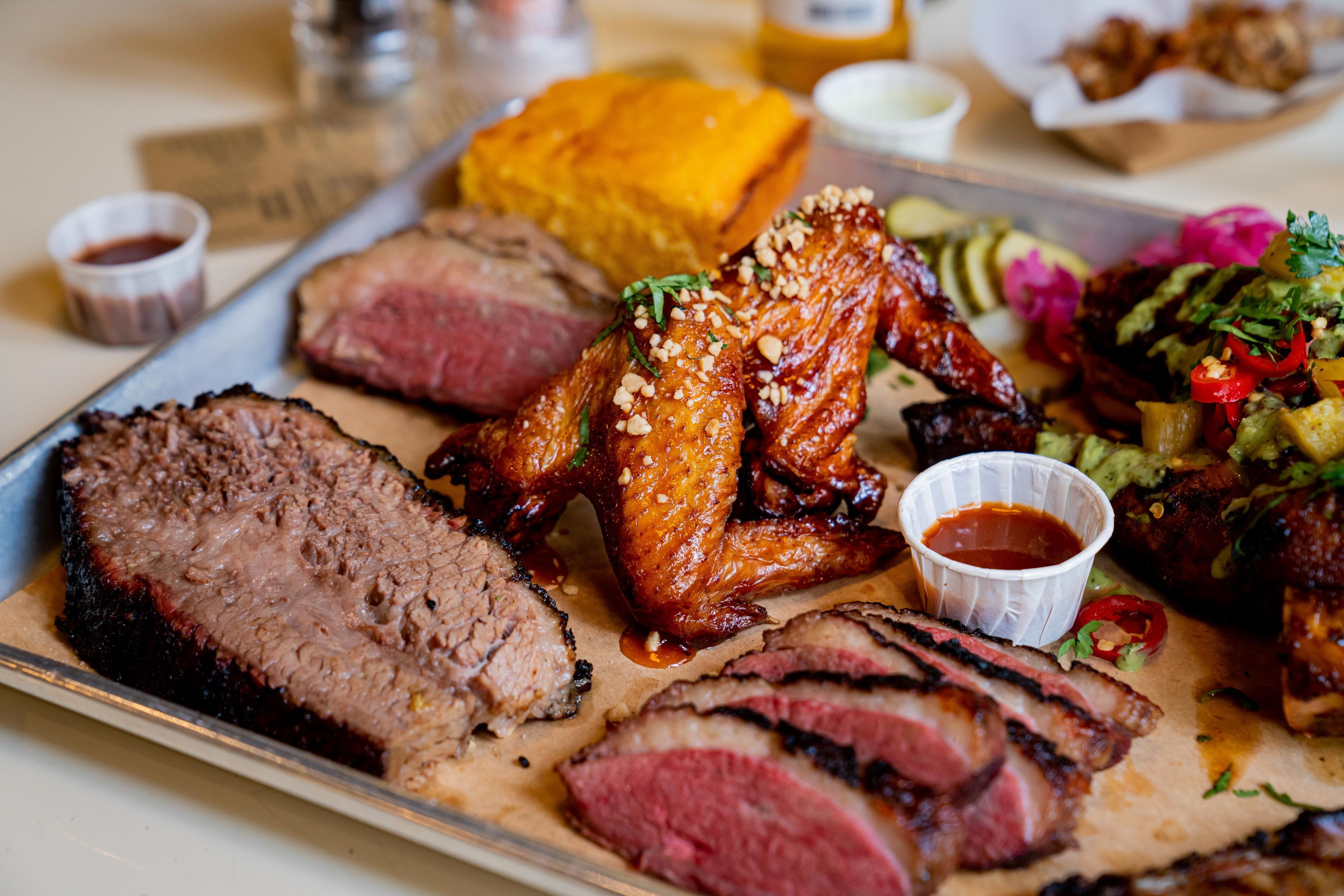 A platter featuring barbecue brisket, wings, and pulled pork, with sides of cornbread and pickles.