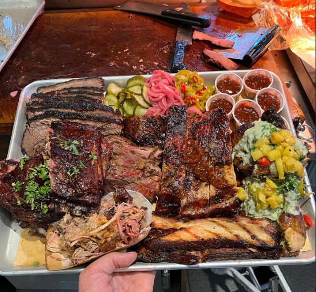 A large platter featuring various barbecue meats, including brisket and pulled pork, alongside pickles and sauce.